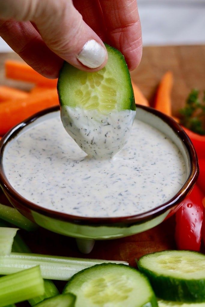 Ranch dressing in a bowl and a hand dipping a cucumber slice